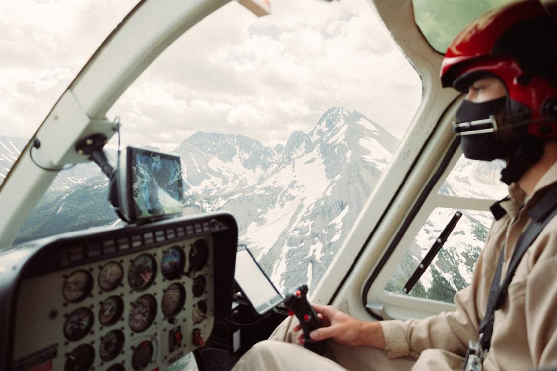 helicopter pilot with a red helmet flying over scenic snowy mountains, with a clear view of the cockpit control panel