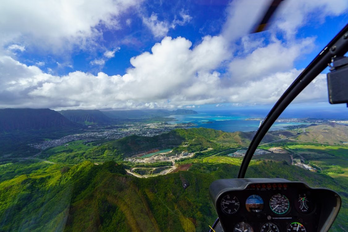 aerial view from a helicopter cockpit featuring bright green hills and bright blue ocean under a cloudy sky