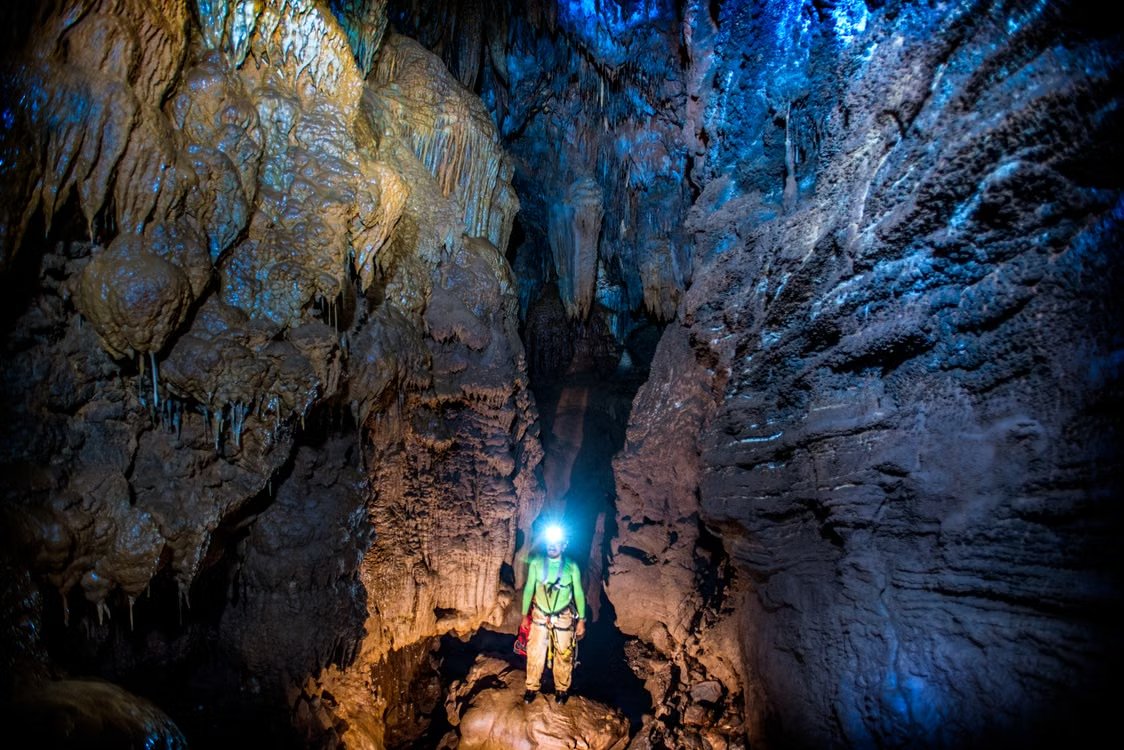 man in brown pants and a neon shirt wearing a helmet light standing in a cave with stalactites in Santiago de Querétaro, Mexico