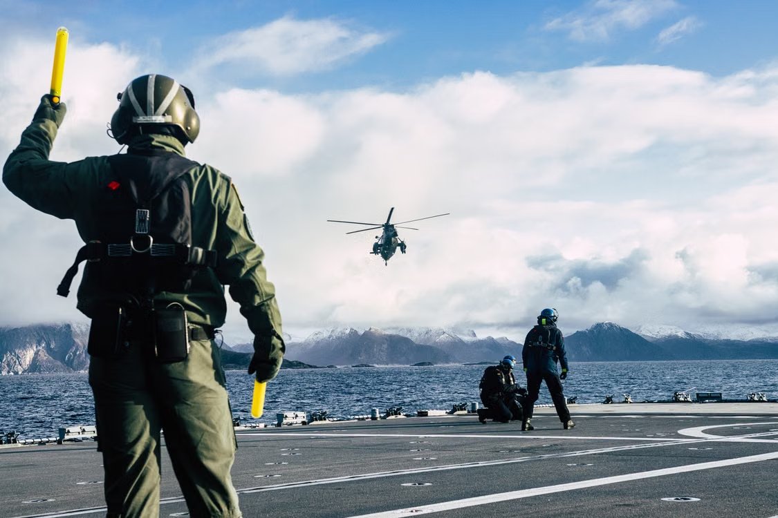 military helicopter flying over water toward an aircraft carrier landing zone guided by ground personnel wearing PPE and waving marshalling wands