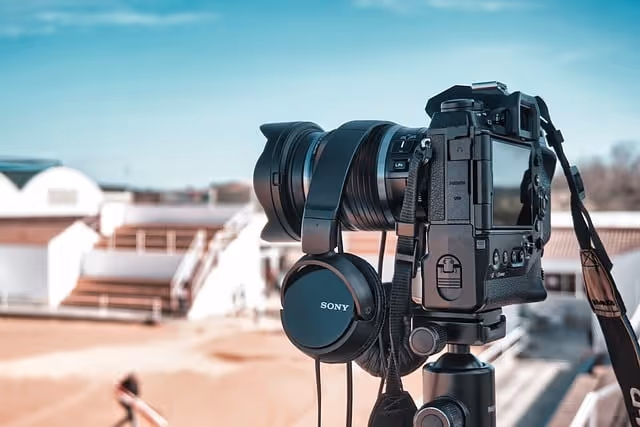 A Sony digital camera with attached lens and headphones mounted on a tripod, photographed against a blurred turquoise sky and rooftop background.