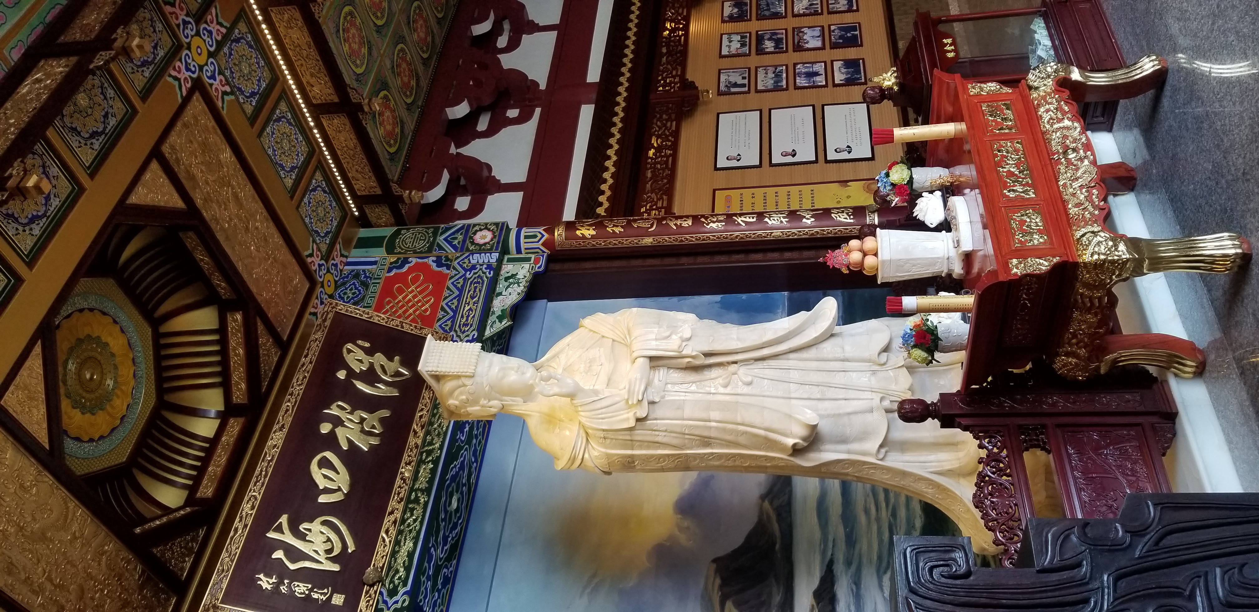 A white Mazu statue beneath an ornate painted temple ceiling