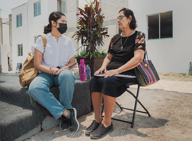 Two women talking to each other, one sitting on a folding stool and another propped up at the end of a low stage. They both have bags and water bottles near and are in front of a newly constructed housing community.