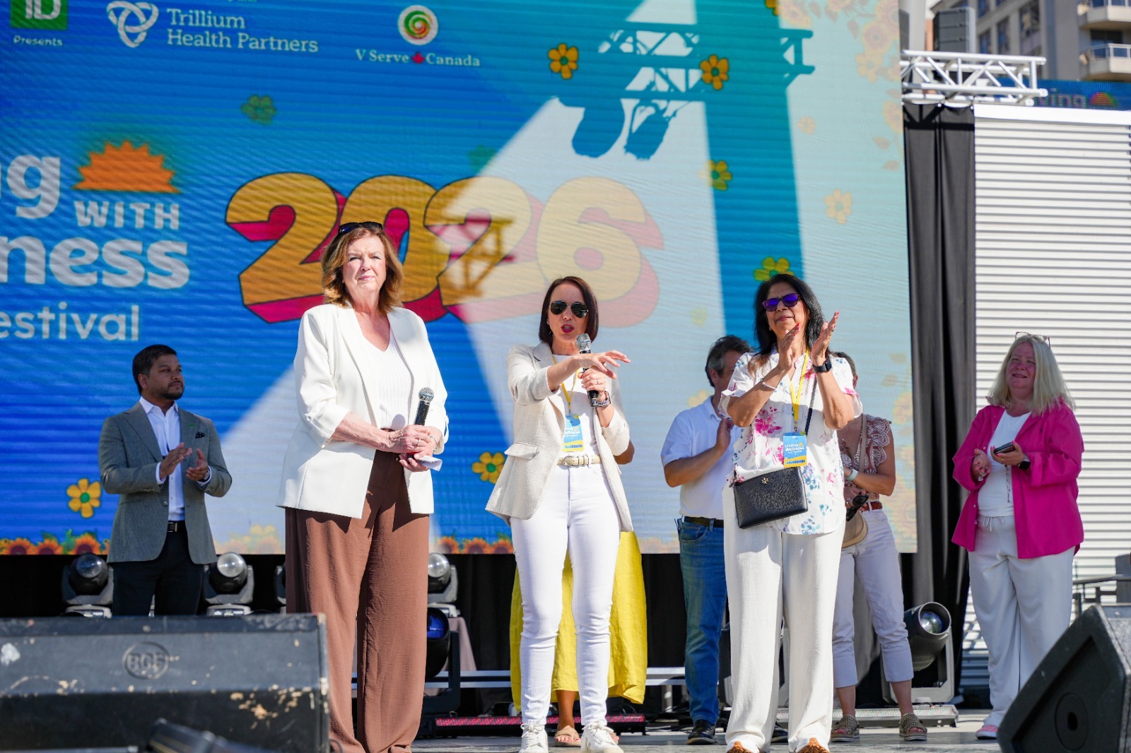Five people smiling and cutting a white cake together that has colorful flowers and the text 'Living with Wellness Arts Festival' written on it, with city buildings visible through large windows behind them.