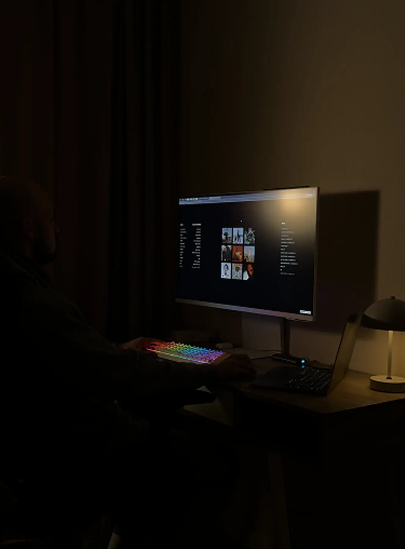 Person sitting in a dark room using a computer with a rainbow-lit keyboard and a large monitor on a desk illuminated by a small lamp.