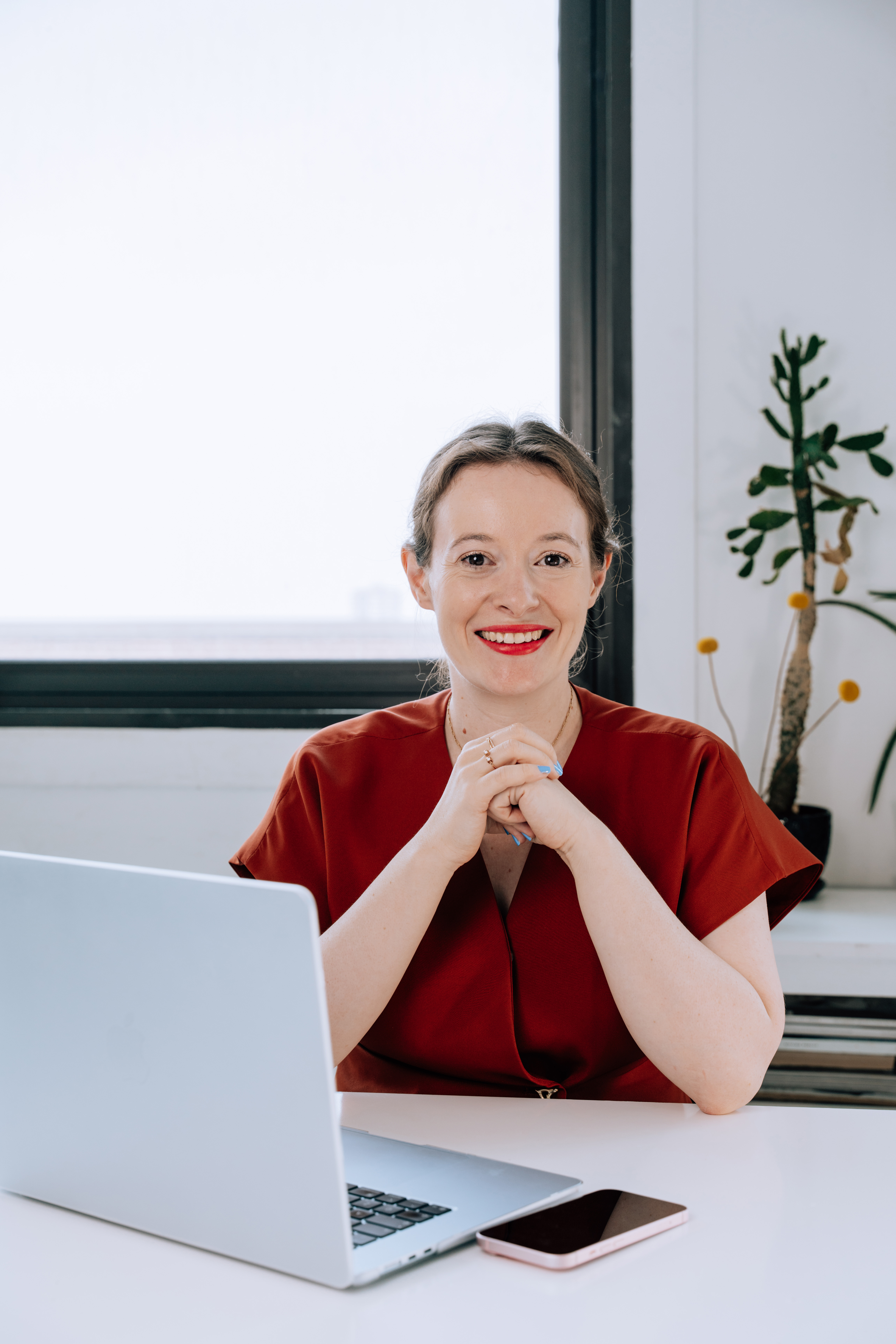 Smiling woman in a red blouse sitting at a white desk with a laptop and smartphone in front of a window and potted plant.