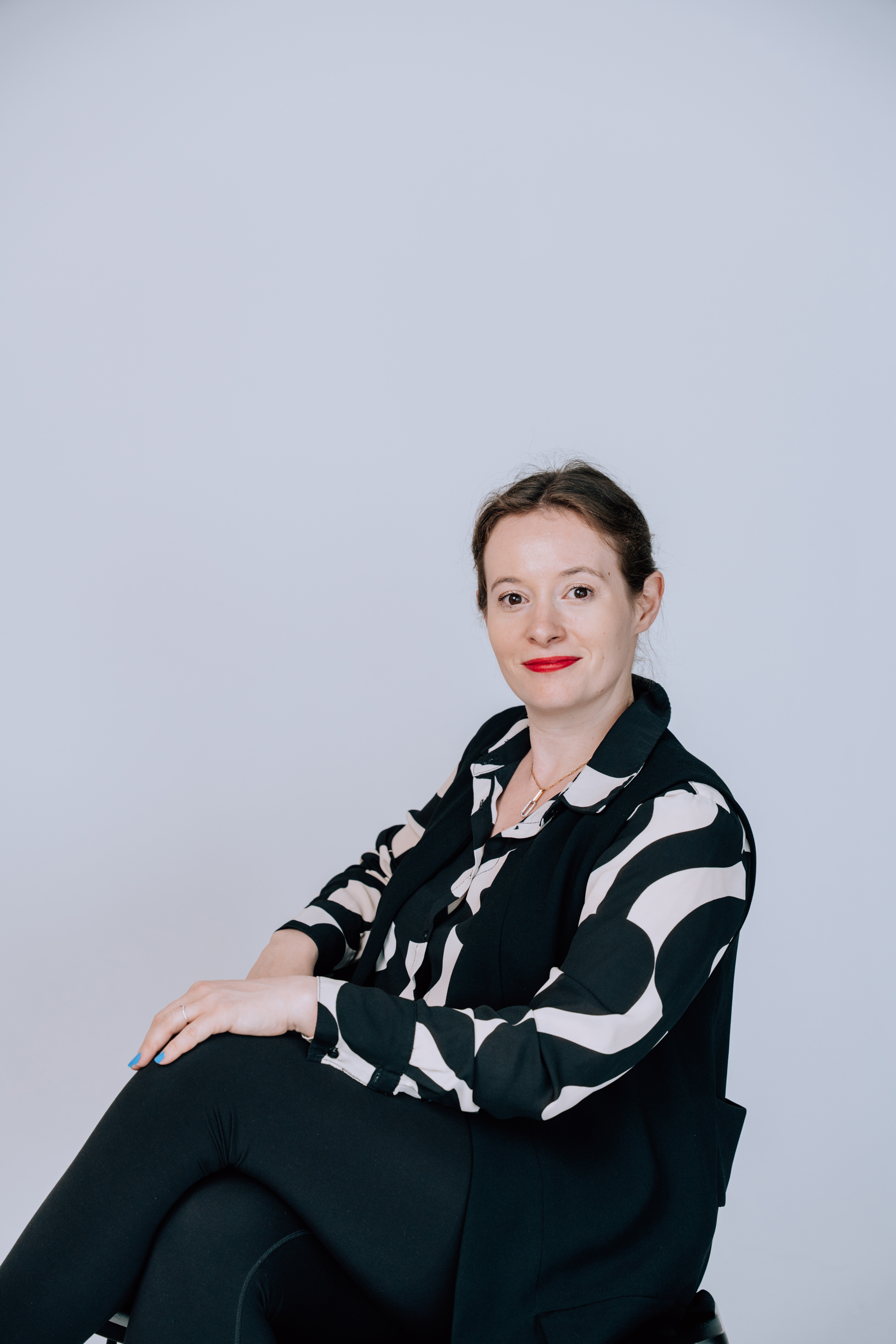 Woman with dark hair tied back, wearing black and white patterned blouse and black vest, seated with legs crossed against a plain light background.