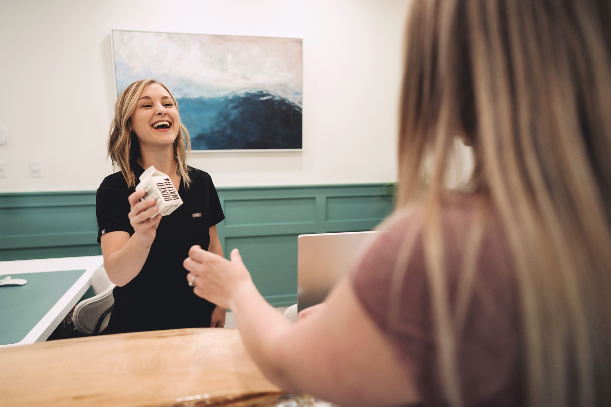 Smiling receptionist handing a small carton of boxed water to a visitor at the front desk.