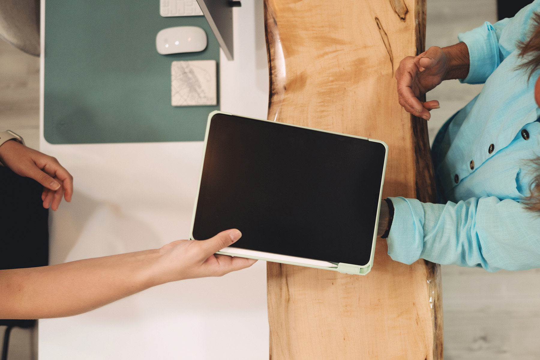 Two people exchanging a tablet over a wooden desk in an office setting.