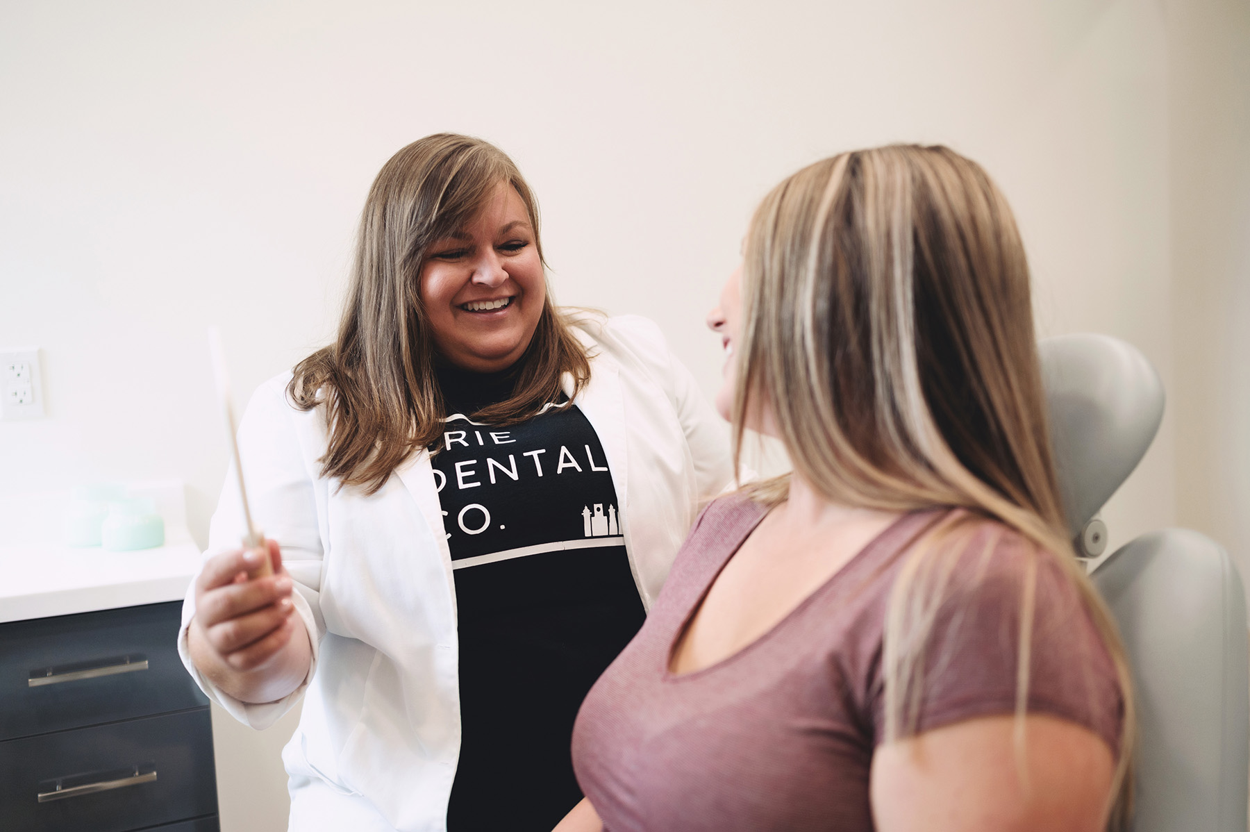 Smiling female dentist in white coat holding a toothbrush, talking to a female patient in a dental chair.