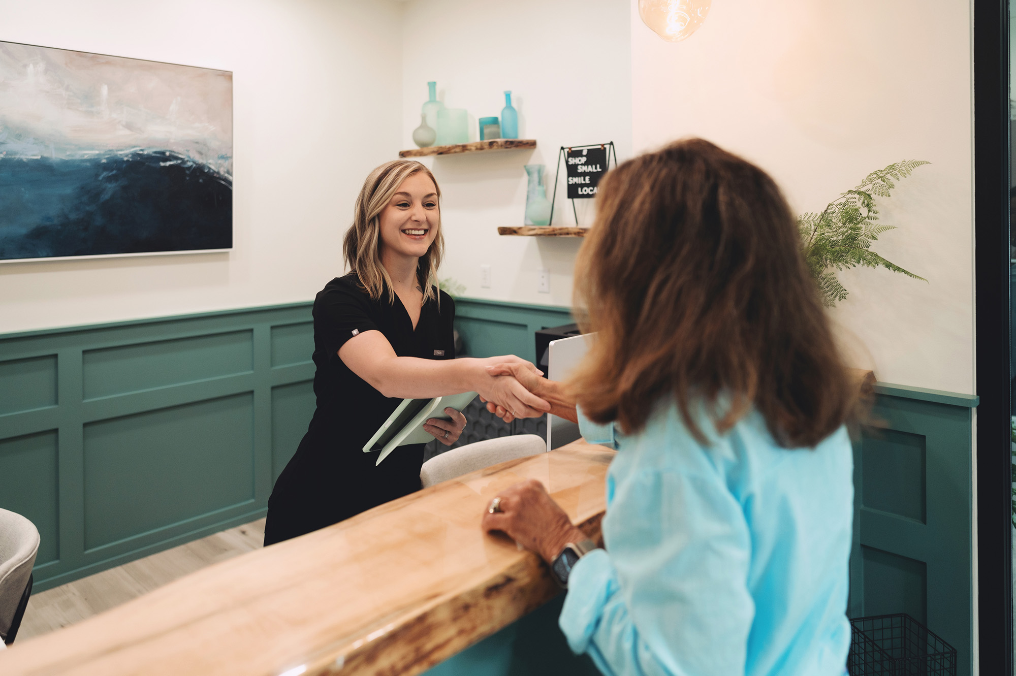 Receptionist in black shaking hands with a visitor in a modern, teal-accented office lobby.