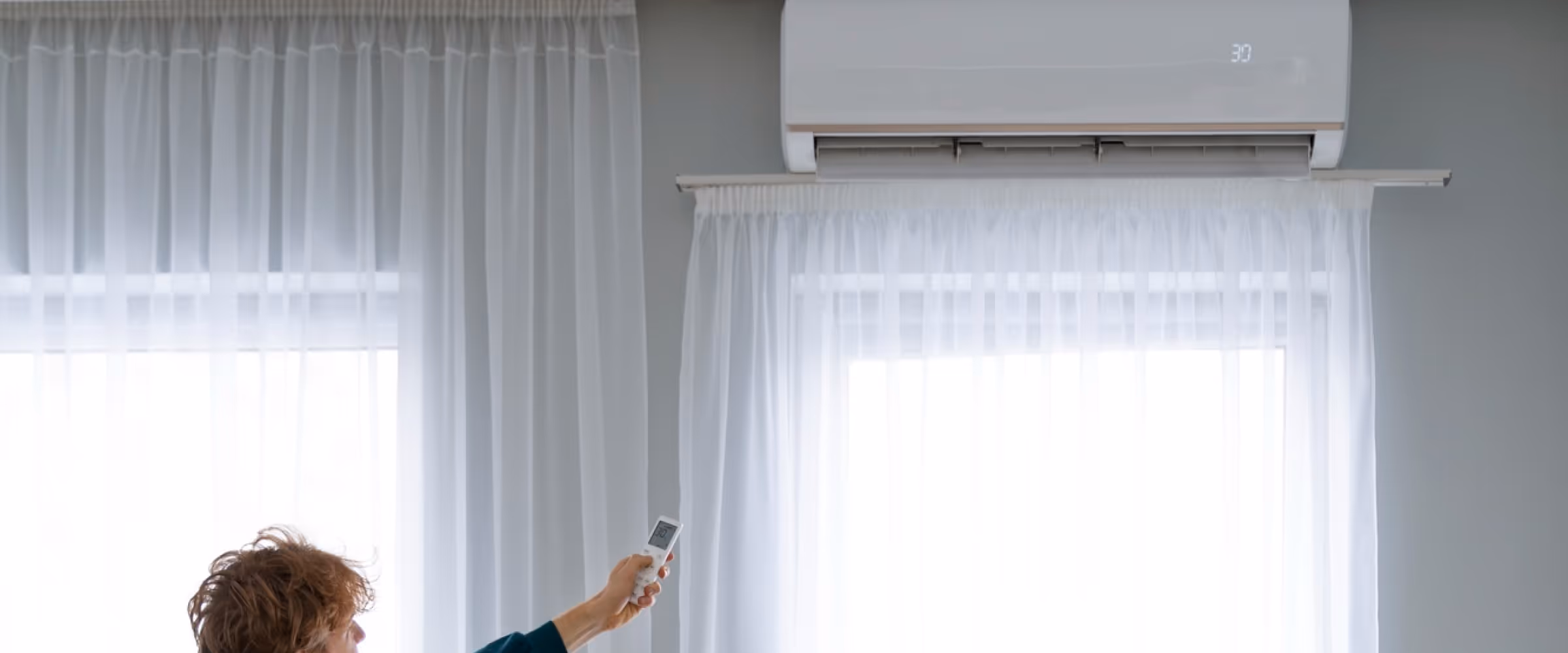 Vrouw richt afstandsbediening op airco aan de muur in een lichte kamer met witte gordijnen.