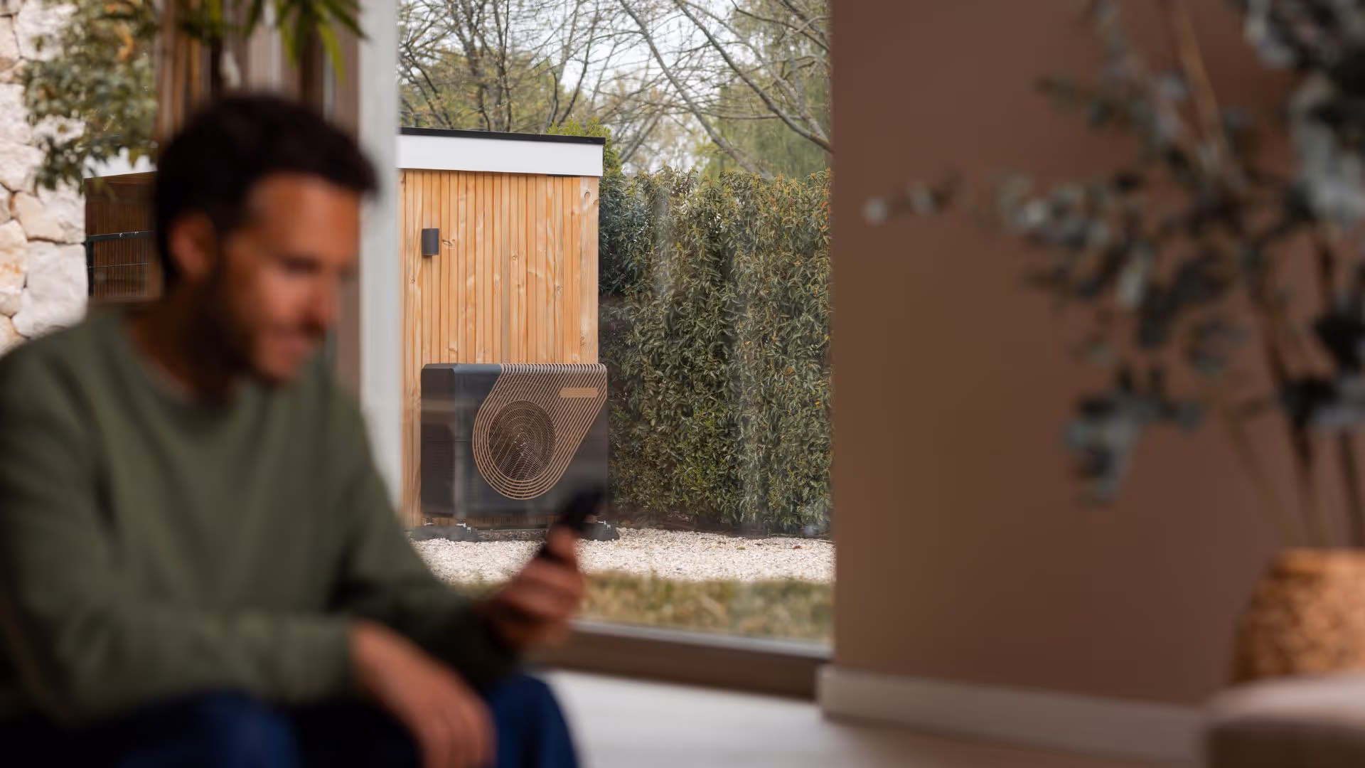 Man in een woonkamer met een warmtepomp op de achtergrond.