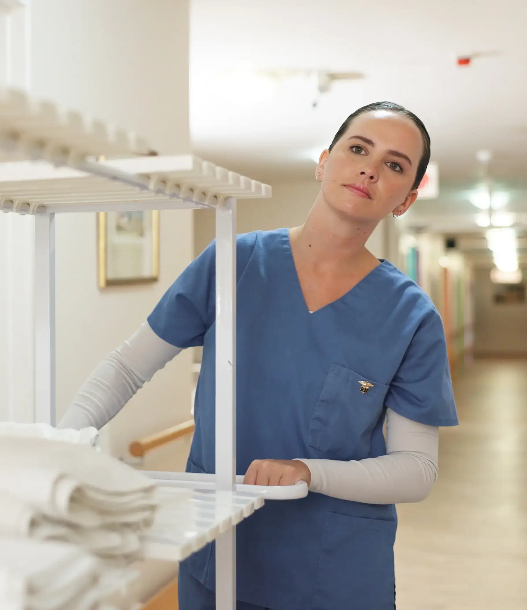 A female nurse in scrubs holds a rack of fresh towels while preparing to change a patient's linens.