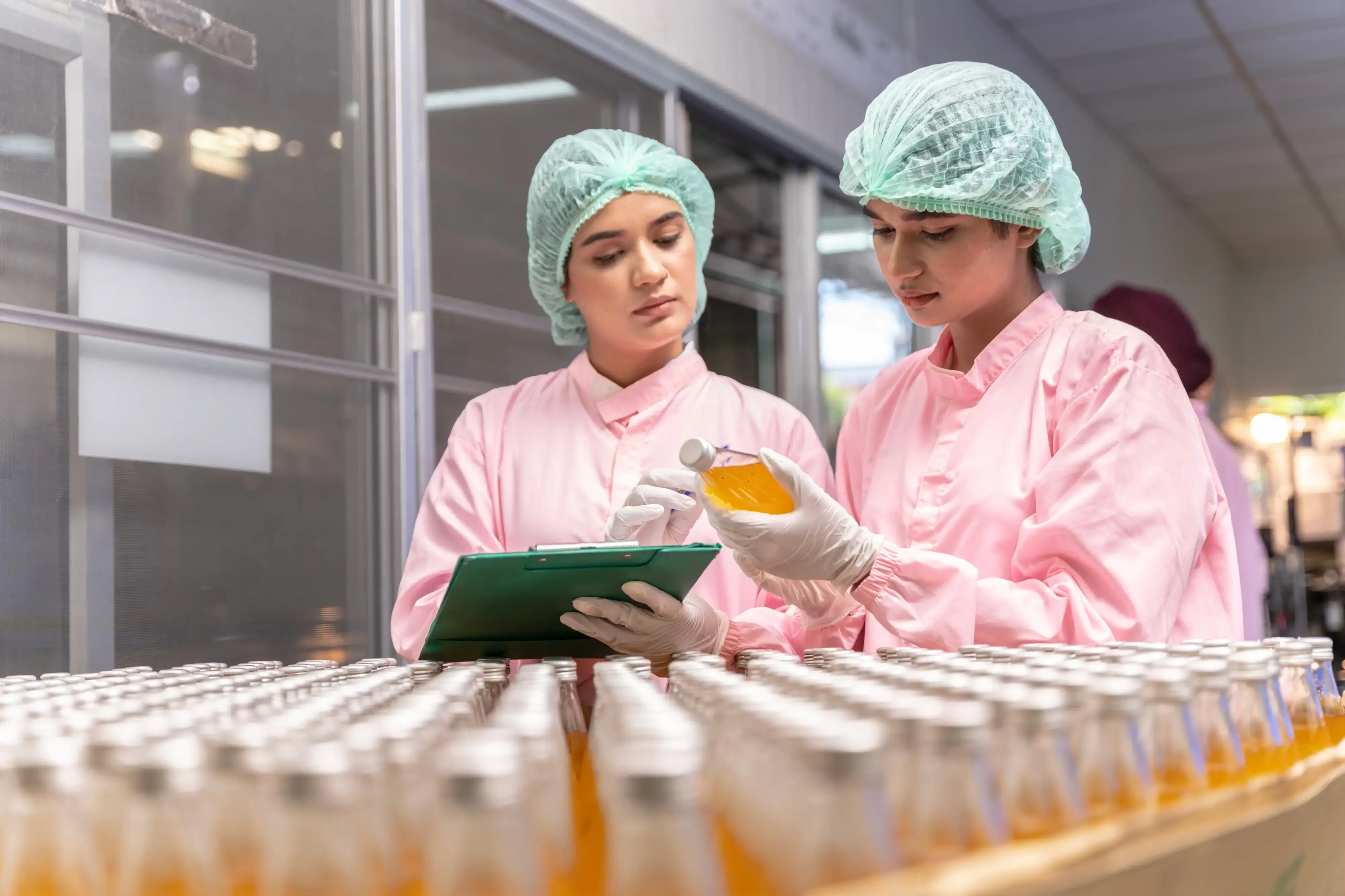 Two women in lab coats examining a tablet while working on a beverage production line.