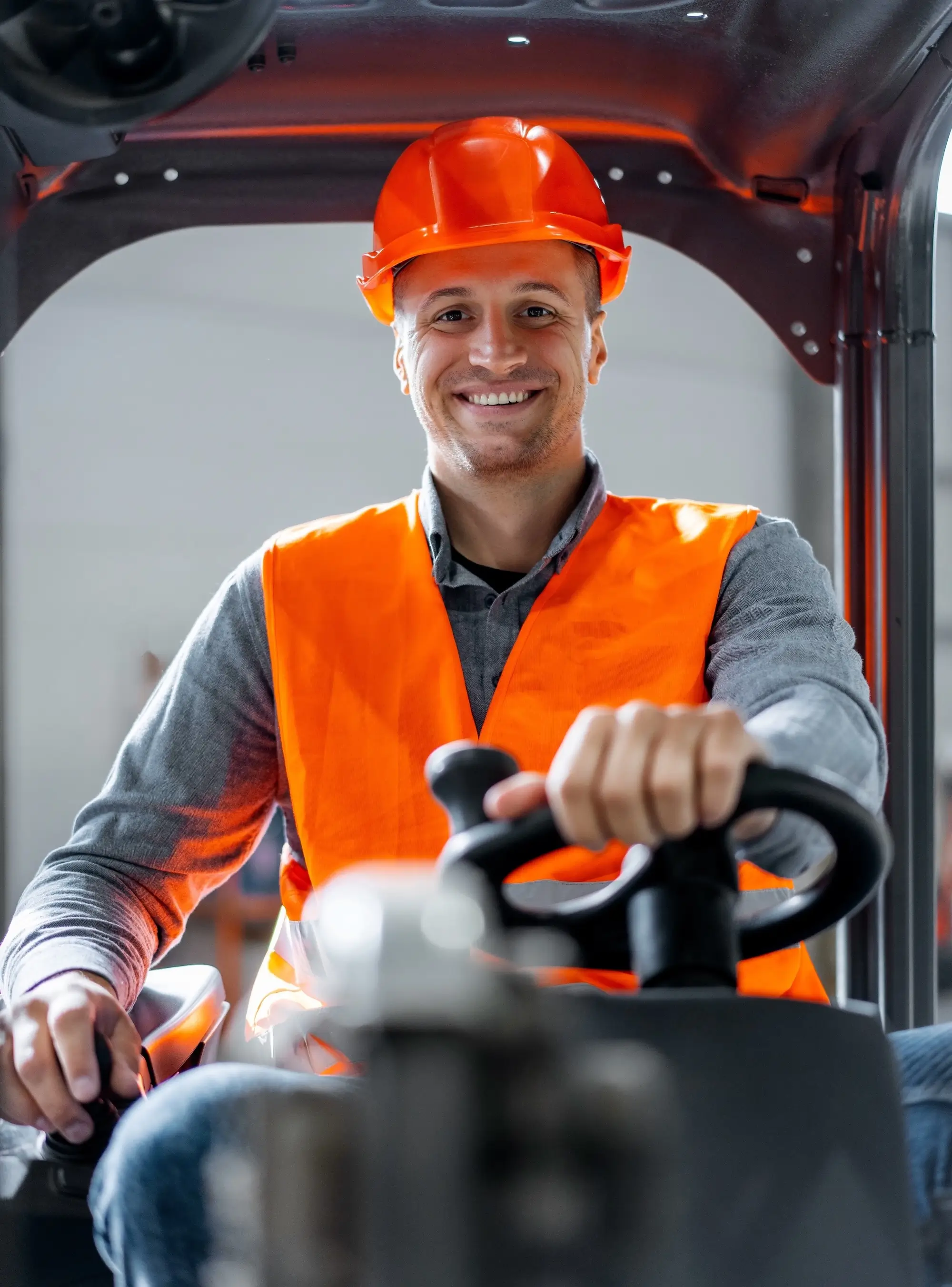 A man wearing an orange vest and hard hat sits in a forklift, focused on his task in a construction environment.