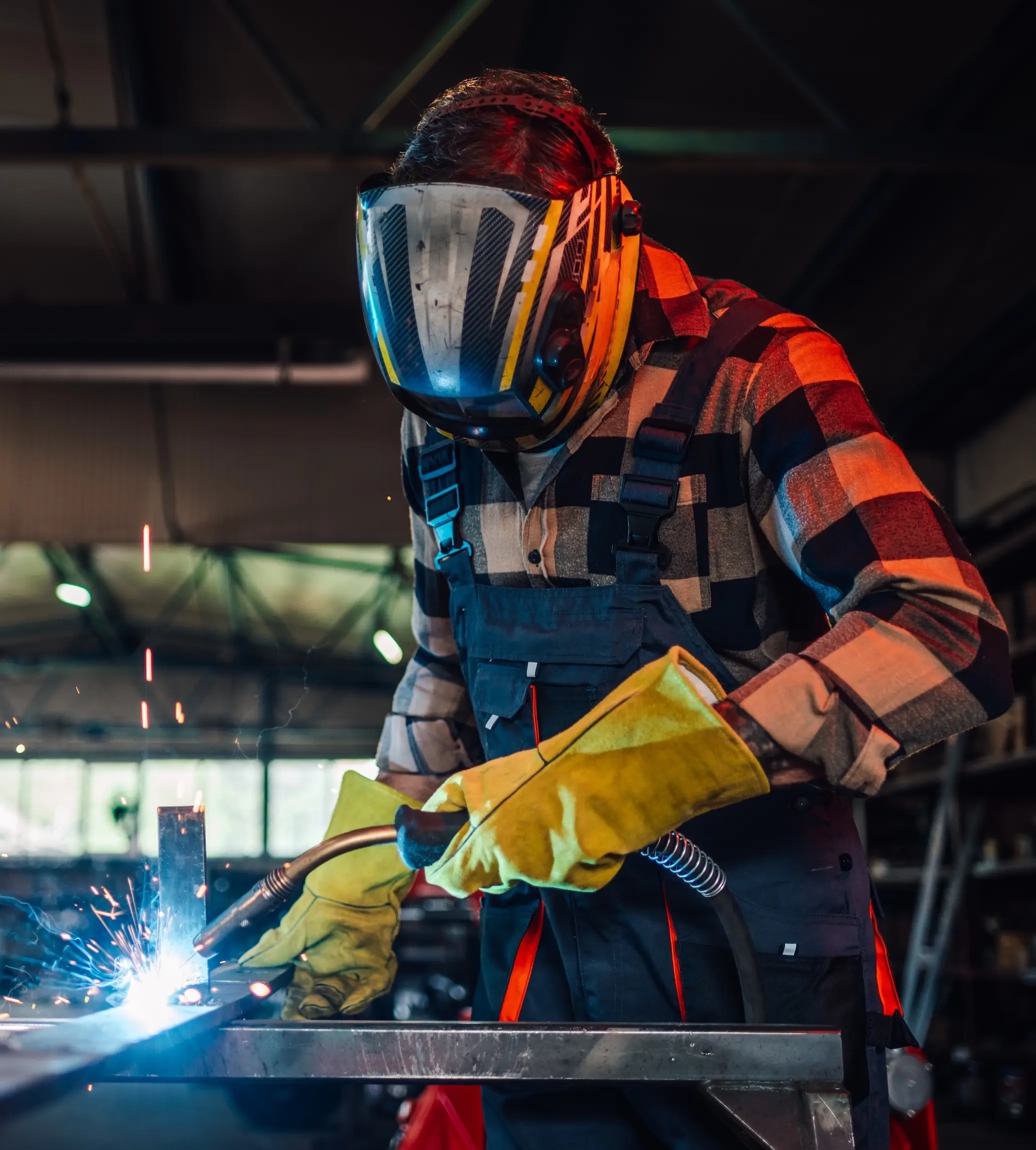 A man in overalls welding metal in a factory, focused on his task with sparks flying around him.