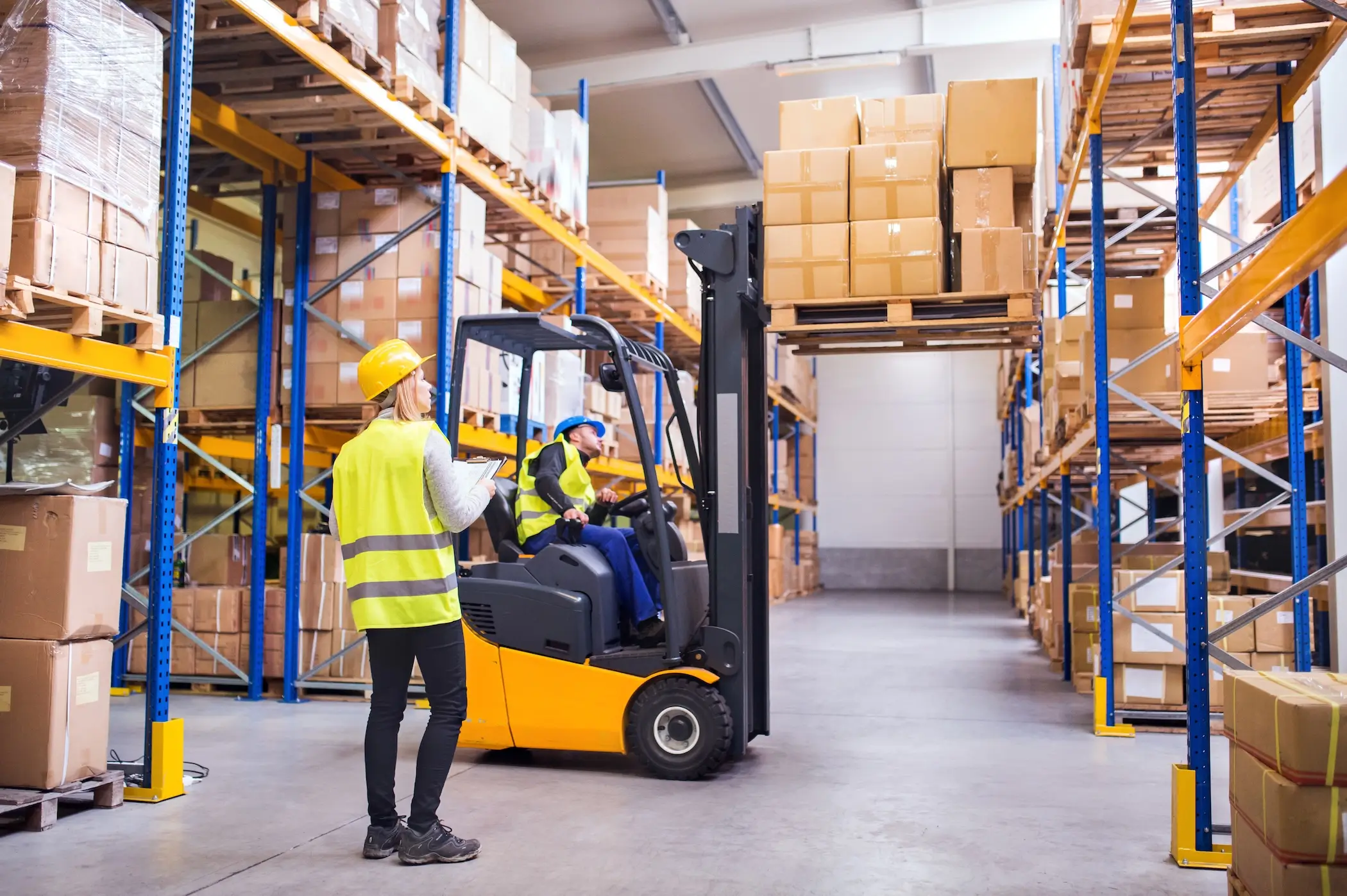 Two warehouse workers operate forklifts among stacked pallets in a busy storage area.