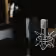 Close-up of a studio microphone with a shock mount and pop filter in a dimly lit recording space.