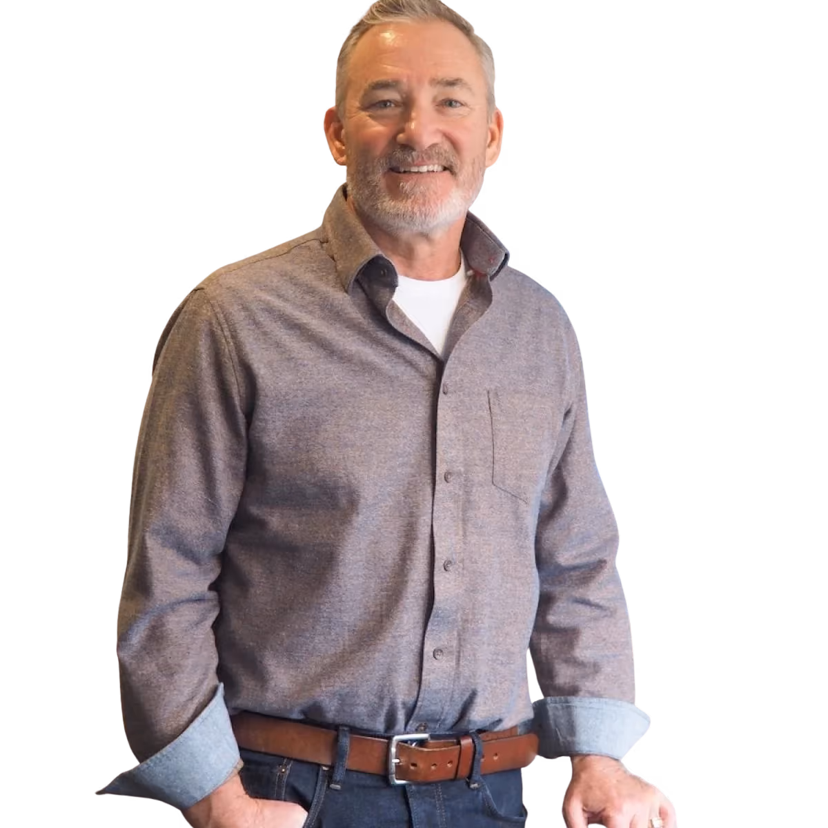 Middle-aged man with gray hair and beard wearing a gray button-up shirt over a white undershirt, dark jeans, and a brown belt, smiling with one hand in his pocket.