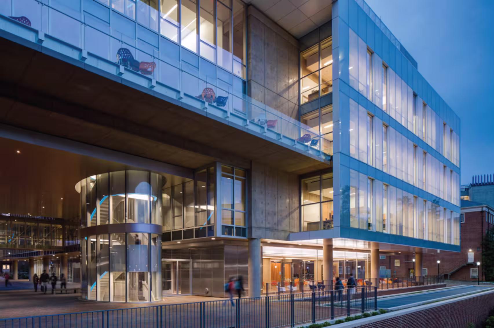 Modern glass-fronted building illuminated at dusk with visible interior spiral staircase and people walking nearby.