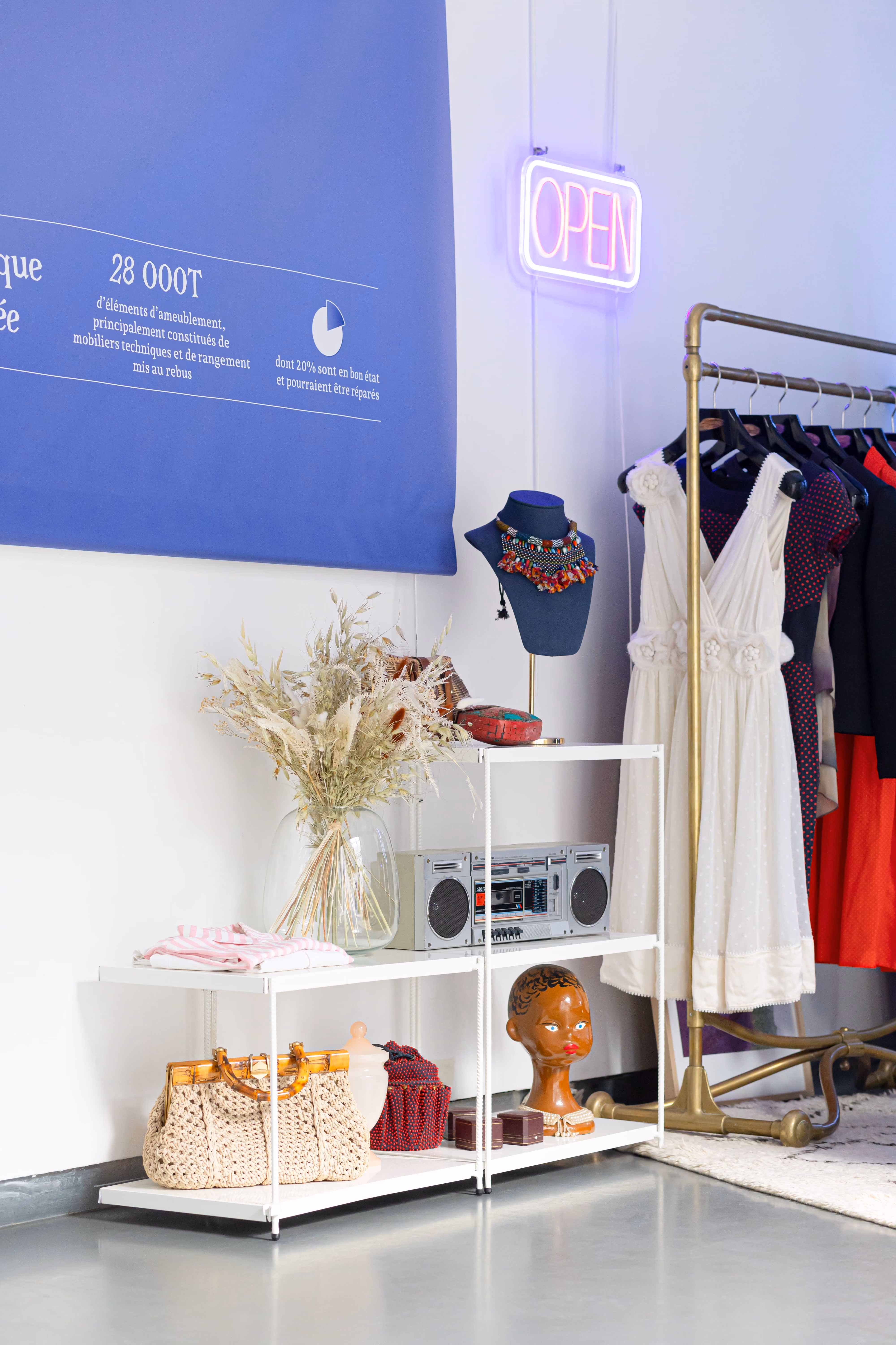Corner de boutique en réemploi : portant de vêtements, étagères chinées et enseigne néon