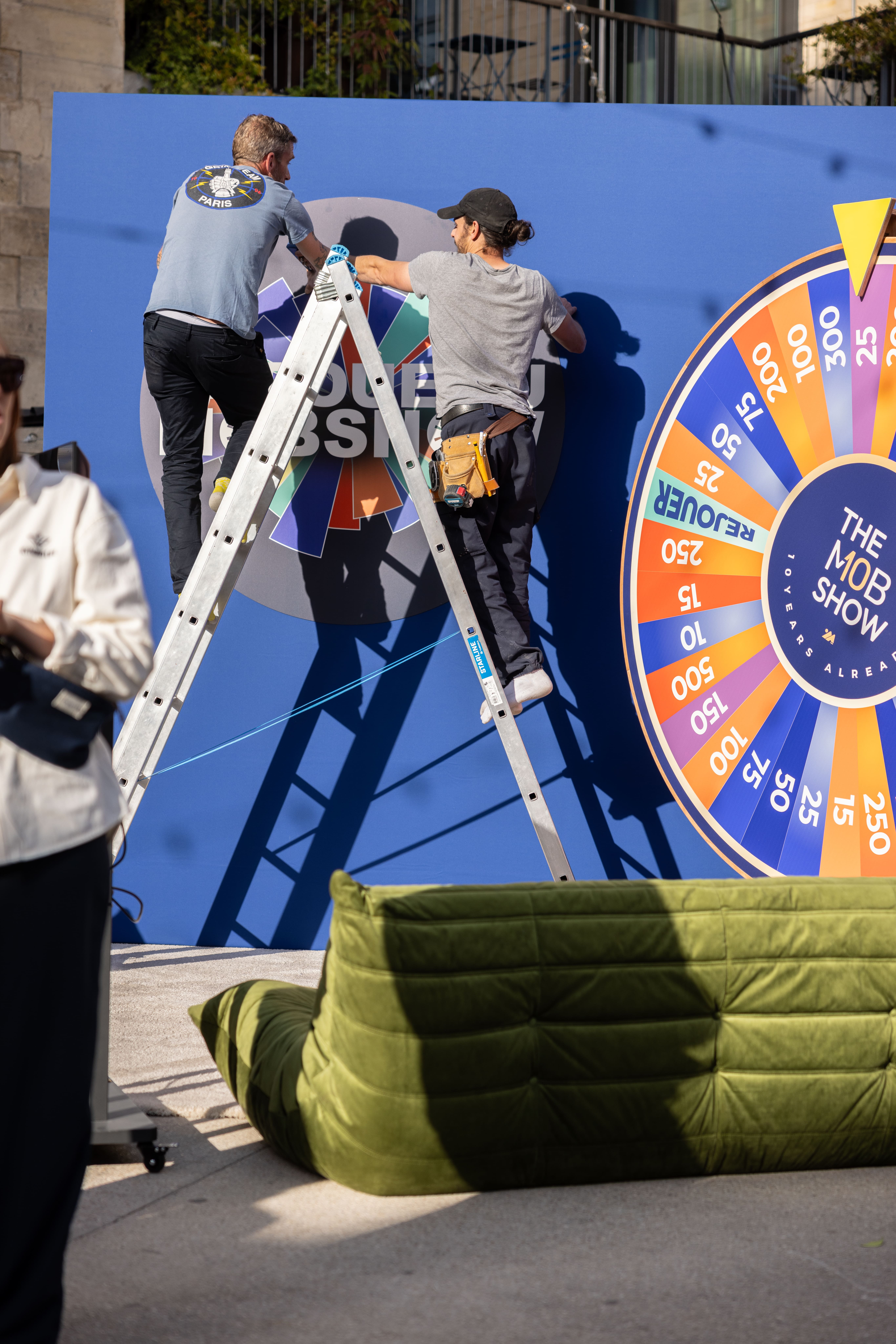 Deux ouvriers sur échelle qui accrochent un logo sur fond bleu