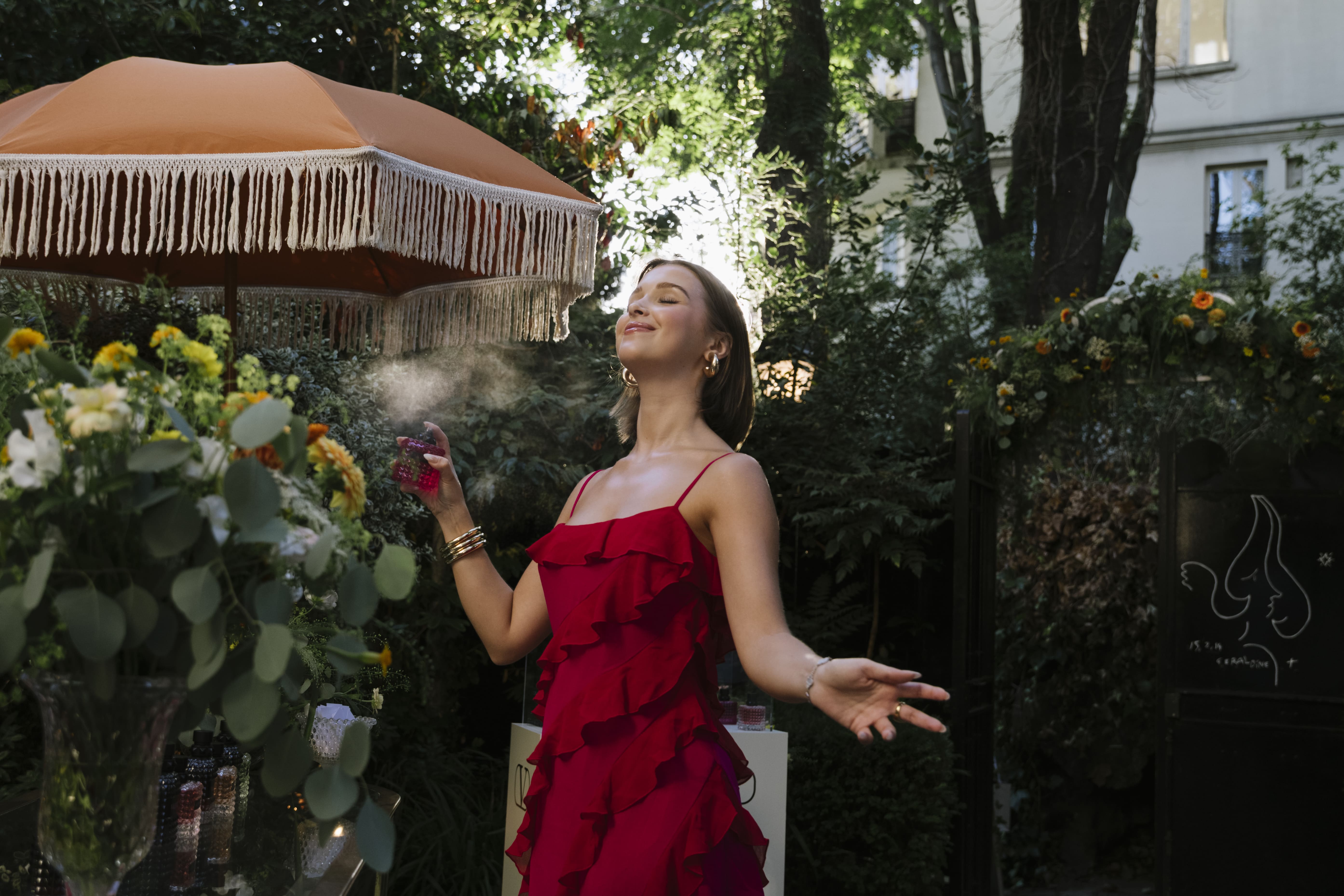 invitée qui se parfume, robe rouge, parasol orange, composition florale, vase, parfums sur présentoirs