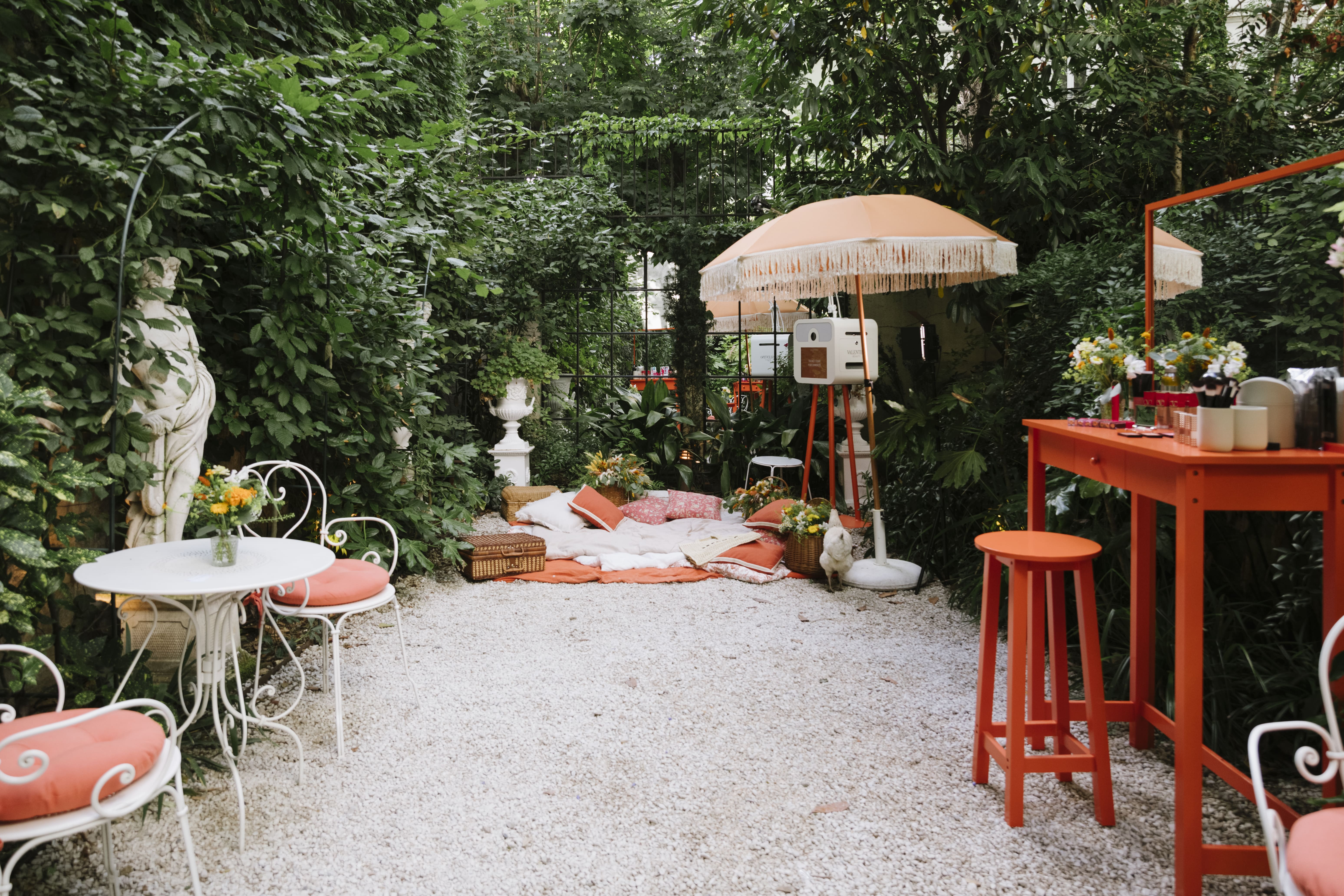 mise en scène du photocall, coiffeuse orange à droite avec maquillage Valentino et parfums, table vintage blanche et chaises assorties, parasol orange à frange, coussins, couvertures beiges, paniers en osier, fleurs