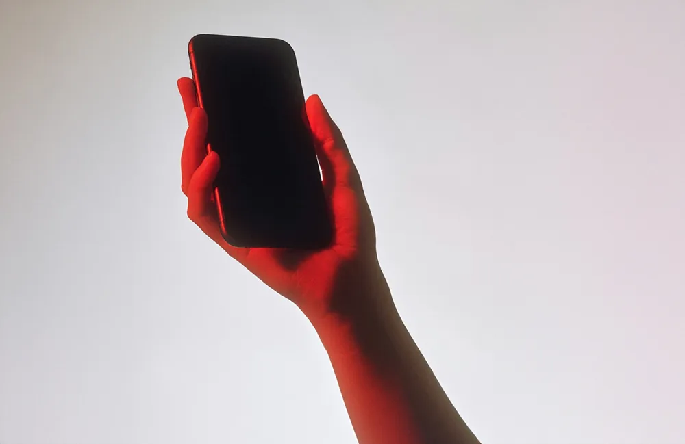 A hand holding up a smartphone with a blank screen, lit in red against a pale background