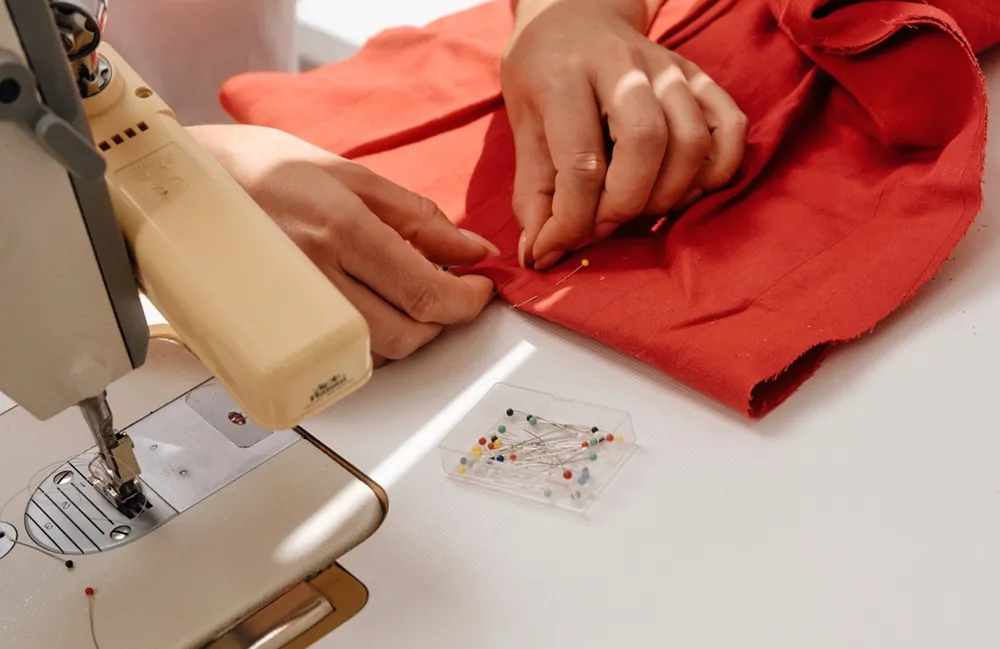 Close-up of a person's hands pinning red fabric at a sewing machine, with a box of coloured pins on the table