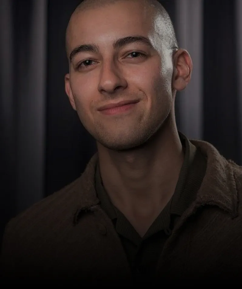 Portrait of a young man with a shaved head, slight smile, and wearing a brown jacket over a dark shirt, against a dark, blurred background.
