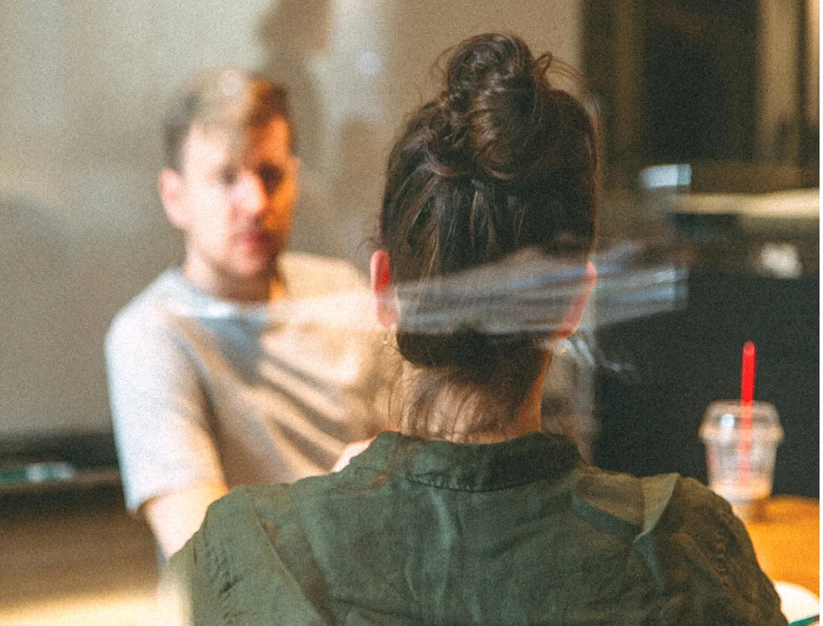 A woman with dark hair tied in a bun wearing a green shirt is sitting across from a man in a light-colored shirt, facing each other in conversation inside a room with a blurred background.
