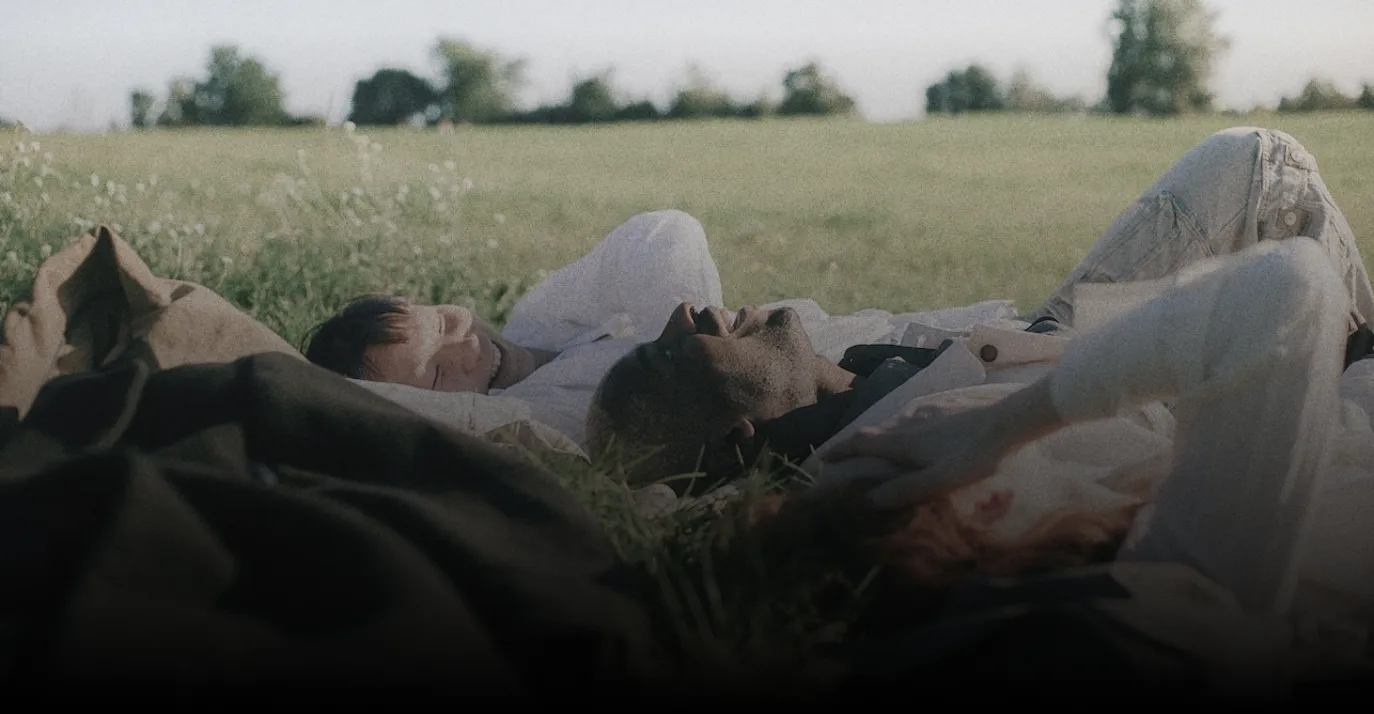 Three people lying in a grassy field under sunlight, appearing relaxed and enjoying each other's company.