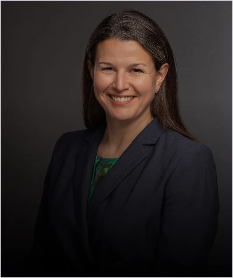 Smiling woman with straight brown hair wearing a dark blazer and patterned top against a dark background.