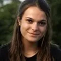 Portrait of a young woman with long brown hair and a slight smile, wearing a black top, outdoors with blurred greenery in the background.