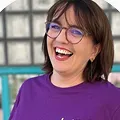 Smiling woman with brown hair wearing glasses and a purple shirt, standing outdoors with a blurred background.