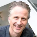 Smiling middle-aged man with light skin and short, light brown hair wearing a dark-colored shirt.