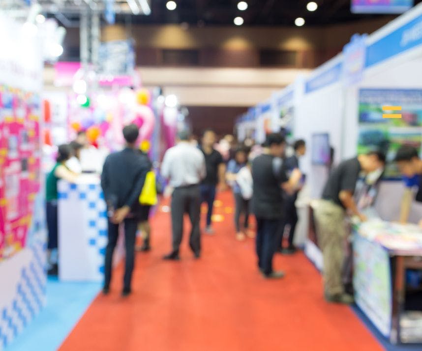 Blurry shot of trade show attendees and exhibitors in the aisle of a trade show with an orange carpet and several trade show booths