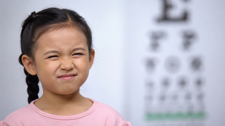 Child squinting during an eye test with a blurred eye chart in the background