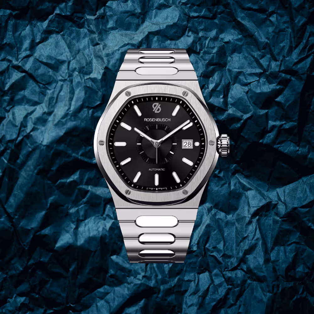 Rosenbusch-Modell „The Quest“ mit Edelstahlarmband auf strukturierter, blauer Oberfläche – kraftvolles Produktvisual.