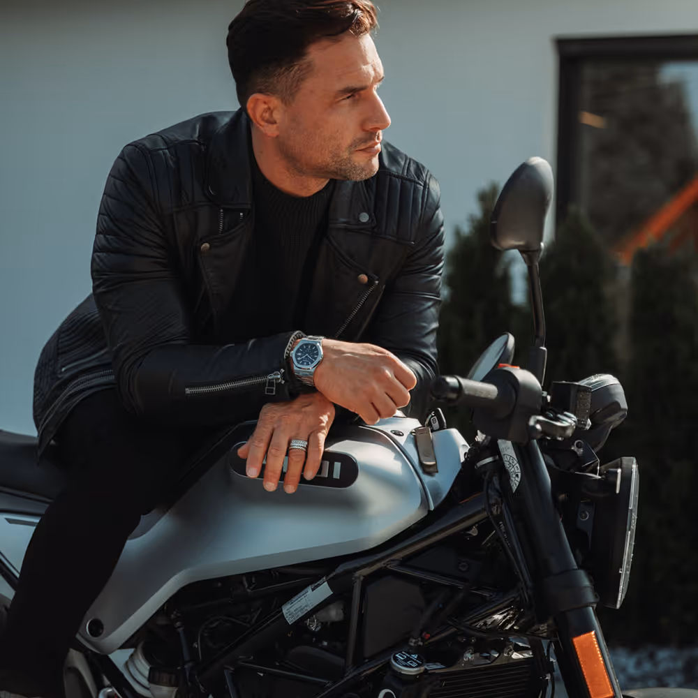 Mann mit Lederjacke und Rosenbusch-Uhr sitzt lässig auf einem Motorrad – ein Statement für Stil und Unabhängigkeit.