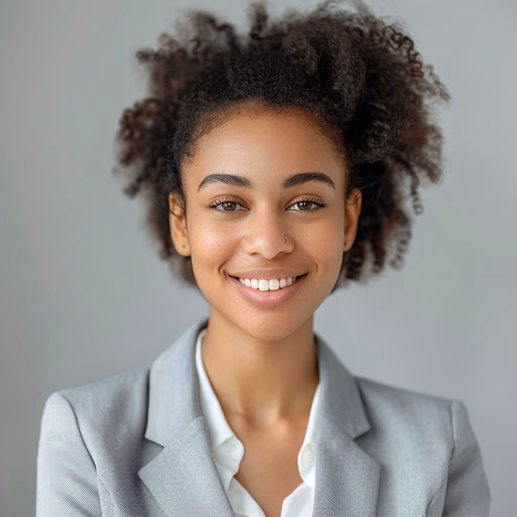 Smiling young woman with curly hair wearing a light gray blazer and white shirt against a light gray background.
