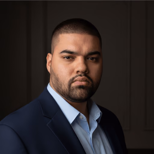 Portrait of a serious man with short hair and beard wearing a dark suit jacket and light blue shirt against a dark background.