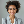 Smiling young woman with curly hair wearing a light gray blazer and white shirt against a light gray background.