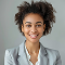 Smiling young woman with curly hair wearing a light gray blazer and white shirt against a light gray background.