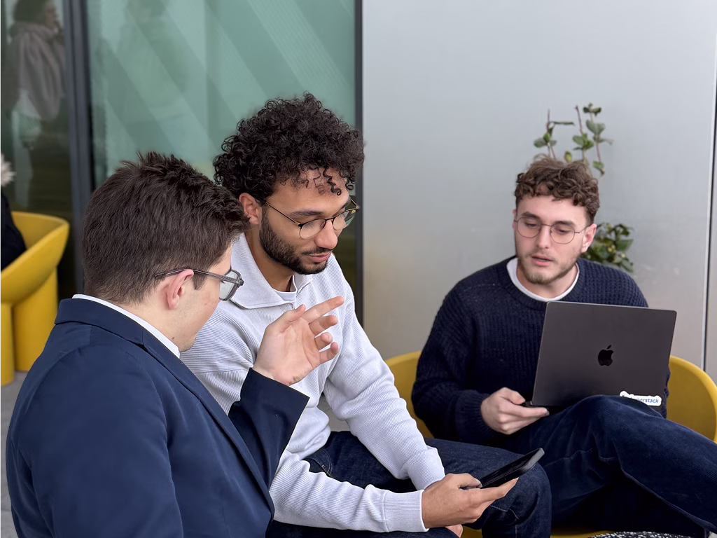 Trois hommes discutent, deux regardent un téléphone, un autre tient un ordinateur portable Apple.