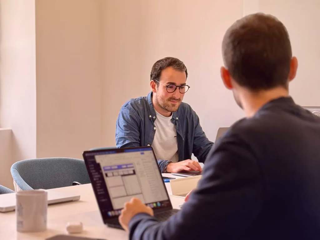 Deux hommes discutant à une table de travail avec un ordinateur portable et une tasse.