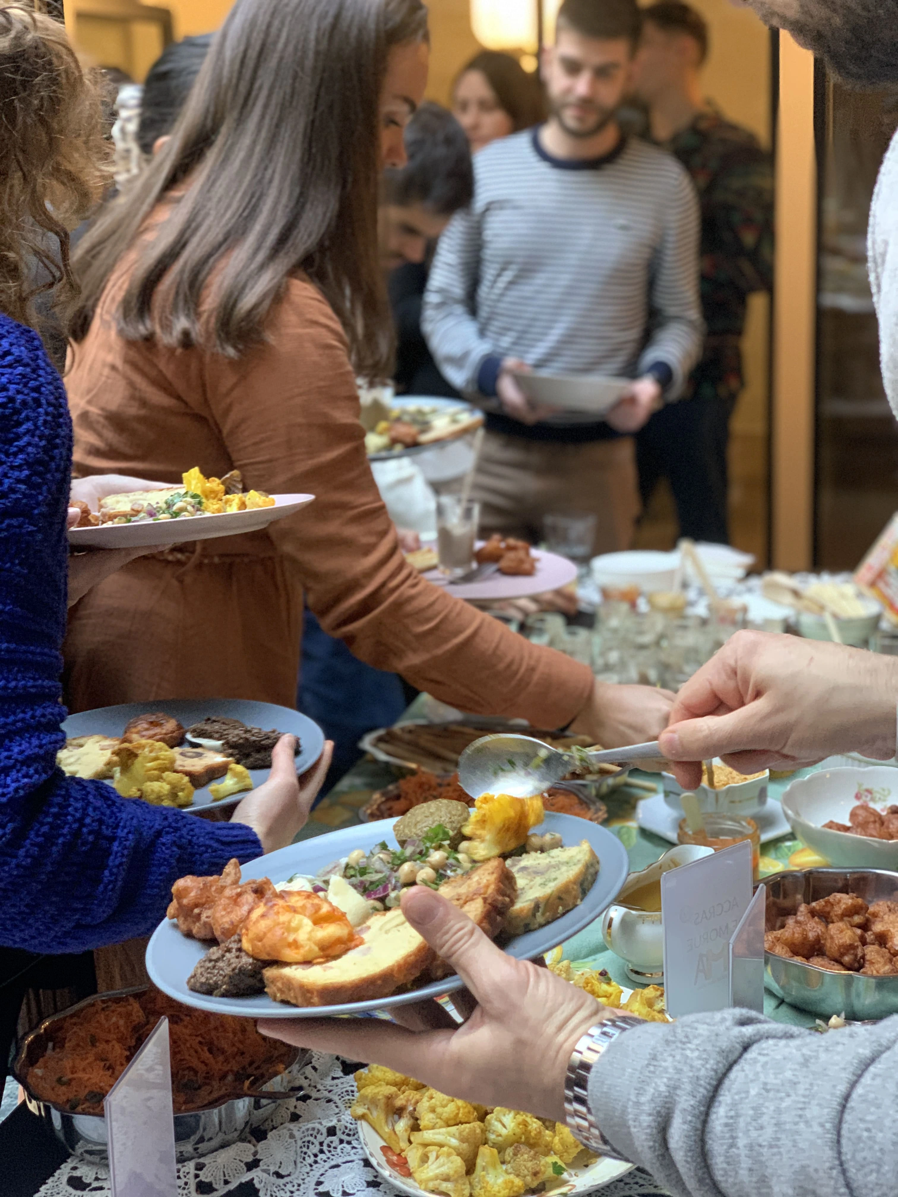 People serving themselves various dishes from a buffet table filled with salads, fried snacks, and breads.