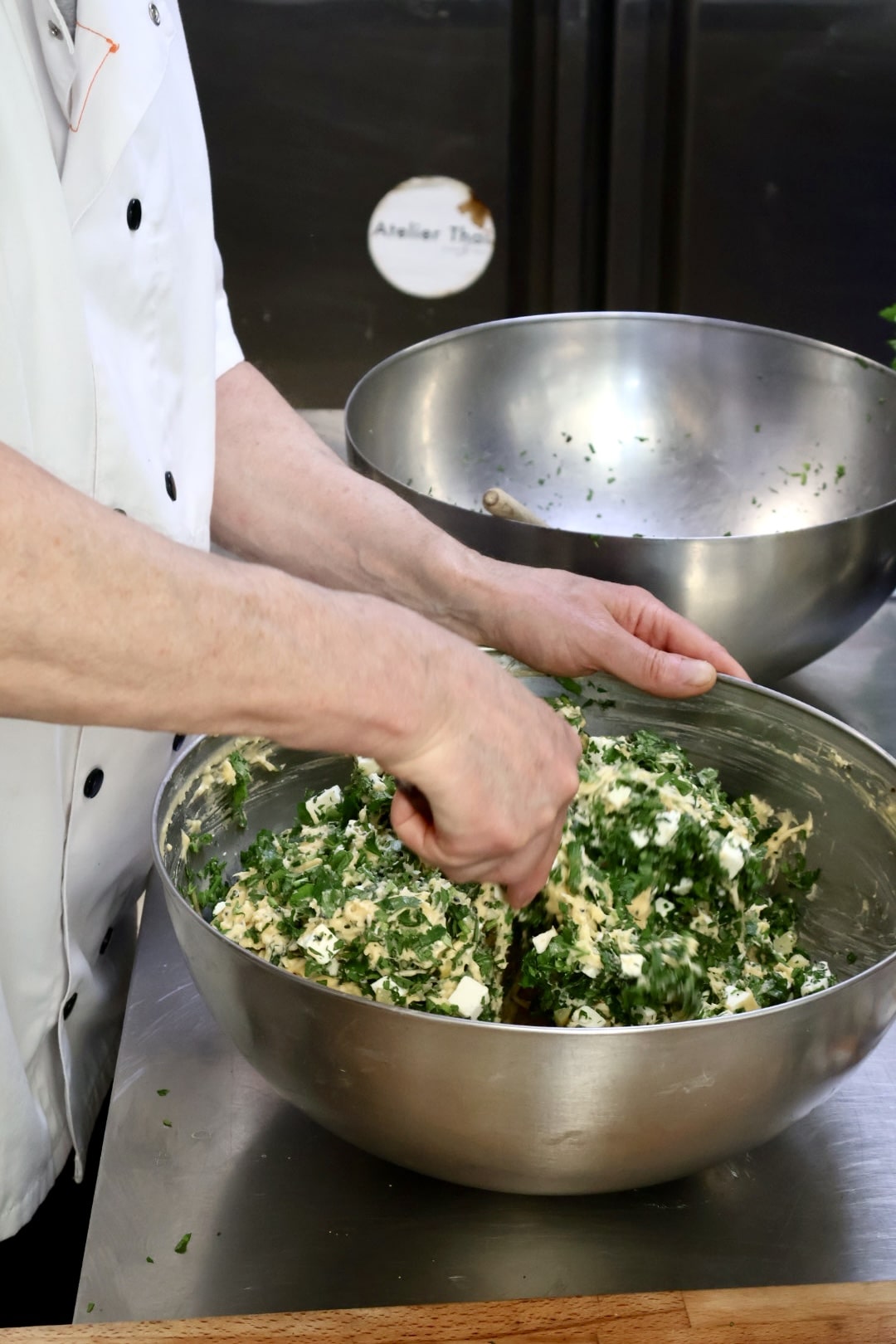 Person in a white chef coat mixing chopped herbs and cheese in a large metal bowl on a kitchen countertop.